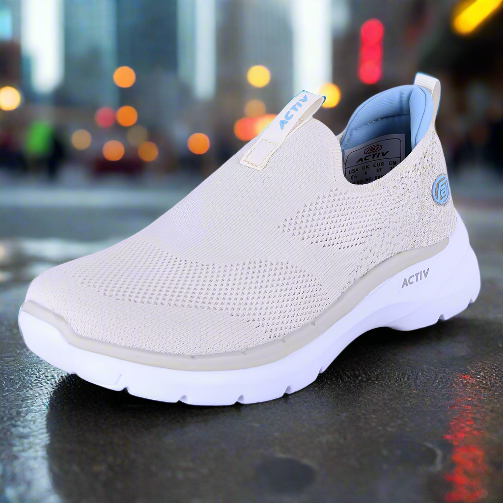 Beige slip-on shoe with white sole on a reflective surface with blurred city lights in the background