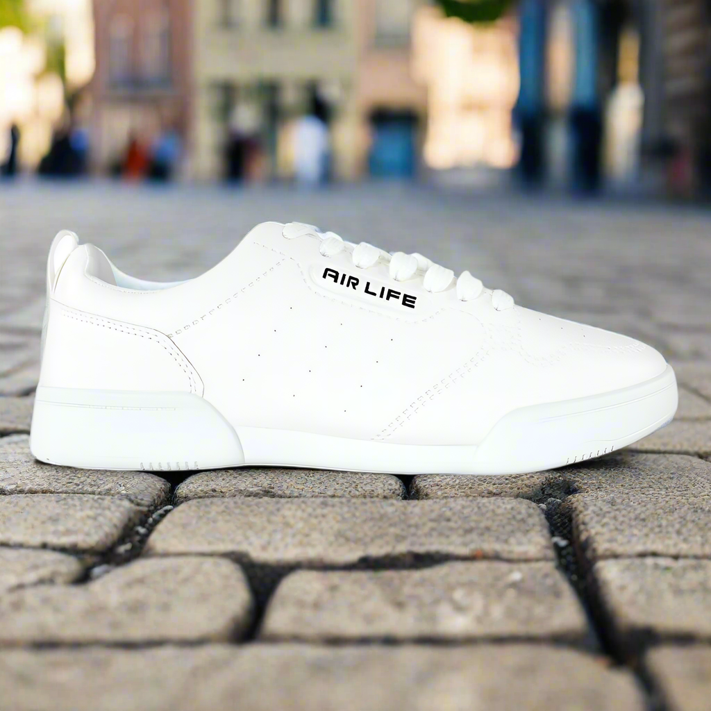 A single white AIRLIFE fashion shoe placed on a cobblestone surface with a blurred background of a street scene.